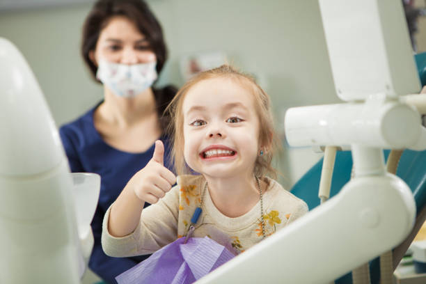 Cheerful kid with broad smile after painless teeth polishing procedure sits in comfortable chair at modern dentist office with mother and doctor in ask who stand behind.